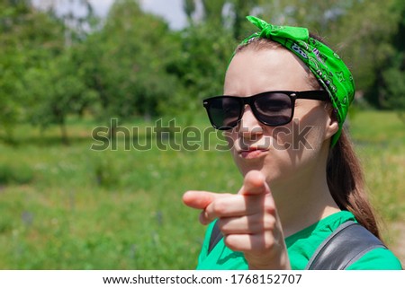 Portrait of a pretty hipster girl in sunglasses, green shirt, bandana standing on nature background on sunny day. Serious emotional caucasian young lady smiles, shows a finger and looks at the camera.