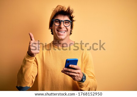 Young handsome man using smartphone wearing glasses over isolated yellow background happy with big smile doing ok sign, thumb up with fingers, excellent sign