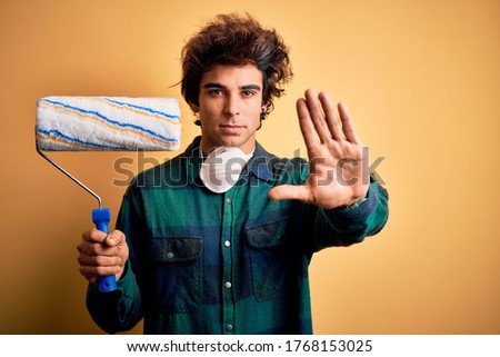 Young handsome painter man holding roller standing over isolated yellow background with open hand doing stop sign with serious and confident expression, defense gesture