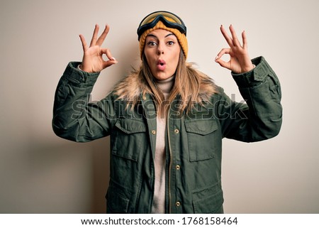 Young brunette skier woman wearing snow clothes and ski goggles over white background looking surprised and shocked doing ok approval symbol with fingers. Crazy expression