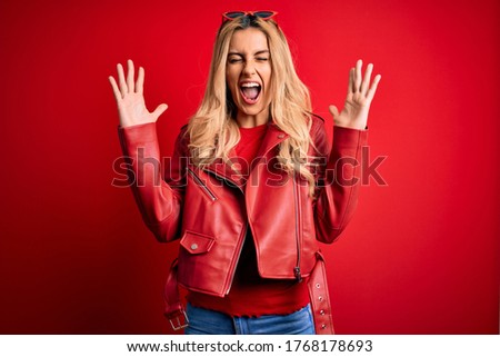 Young beautiful blonde woman wearing casual jacket standing over isolated red background celebrating mad and crazy for success with arms raised and closed eyes screaming excited. Winner concept