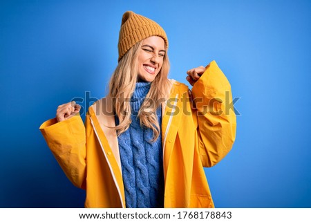 Young beautiful blonde woman wearing raincoat for rainy weather over blue background very happy and excited doing winner gesture with arms raised, smiling and screaming for success. Celebratio.