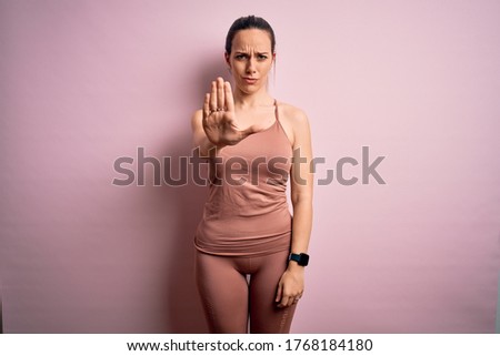 Young blonde fitness woman wearing sport workout clothes over pink isolated background doing stop sing with palm of the hand. Warning expression with negative and serious gesture on the face.