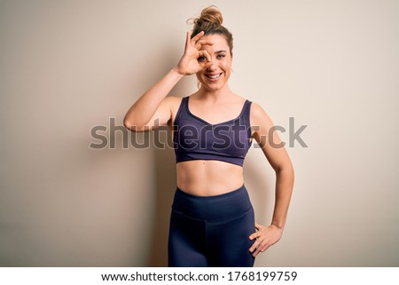 Young beautiful blonde sportswoman doing sport wearing sportswear over white background doing ok gesture with hand smiling, eye looking through fingers with happy face.