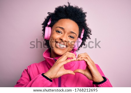Young African American afro woman with curly hair listening to music using pink headphones smiling in love showing heart symbol and shape with hands. Romantic concept.