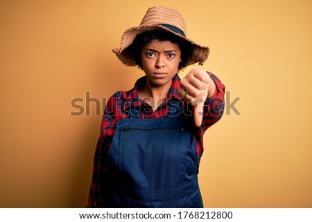 Young African American afro farmer woman with curly hair wearing apron and hat looking unhappy and angry showing rejection and negative with thumbs down gesture. Bad expression.