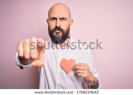 Handsome romantic bald man with beard holding red heart paper over pink background pointing with finger to the camera and to you, hand sign, positive and confident gesture from the front