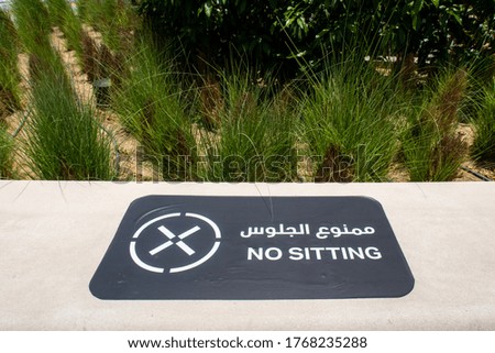 Black NO SITTING placard in english and arabic on stone bench in a park, forbidden to sit due to COVID-19 pandemic, Dubai, United Arab Emirates.