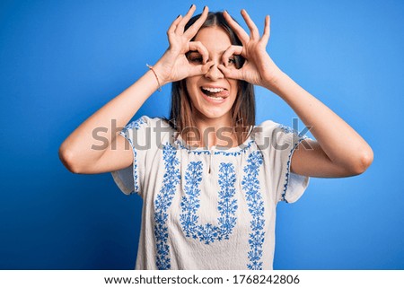 Young beautiful brunette woman wearing casual t-shirt standing over blue background doing ok gesture like binoculars sticking tongue out, eyes looking through fingers. Crazy expression.