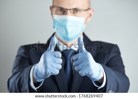 Horizontal shot of Thumbs up sign in focus. A businessman in medical mask and rubber gloves in blur shows thumbs up with both hands.