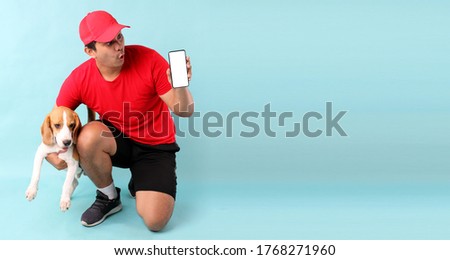 handsome Asian man in red cap and beagle dog,isolated on blue background in studio With copy space. holding mobile phone with blank white empty screen  for ads.