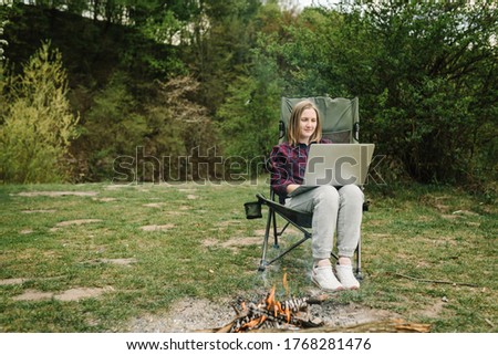 Woman working on laptop online on nature. Young freelancer relaxing in forest. Outdoor activity in summer. Travel, hiking, technology, tourism, people concept. Remote work on a picnic by the fire.