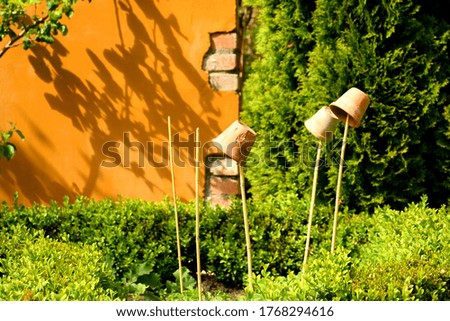 Close up on several clay pots hanging from small wooden sticks located next to an orange brick wall in the middle of a public park seen surrounded with some shrubs and herbs on a hot summer day