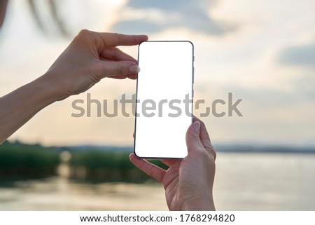 Woman's hands holding a smartphone with white screen mockup on blur background during sunset on the lake. Travel photography taken a memory of the garda lake.