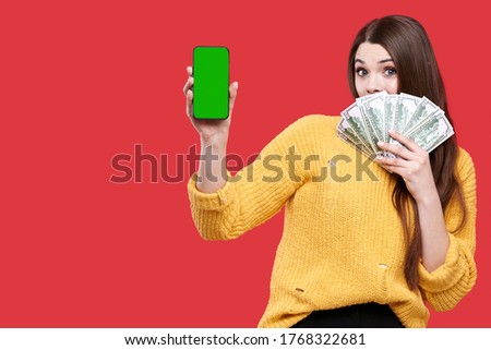 Surprized exciting woman holding mobile phone or smartphone with blank green screen and dollar banknotes, female with money, girl playing bet online casino, isolated on red