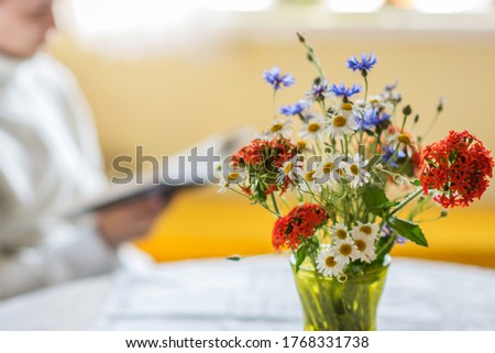 Wild flowers bouquet in the green glass vase on the table with white tablecloth in the natural light, silhouette of girl with book in the background, resting in summer day