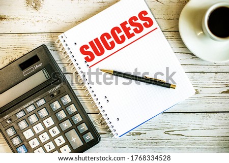 The word SUCCESS is written in red on a notebook on a light table near a calculator and coffee