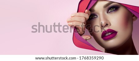 The face of a young beautiful girl with bright makeup and with puffy pink lips looks into the cutout of pink paper.