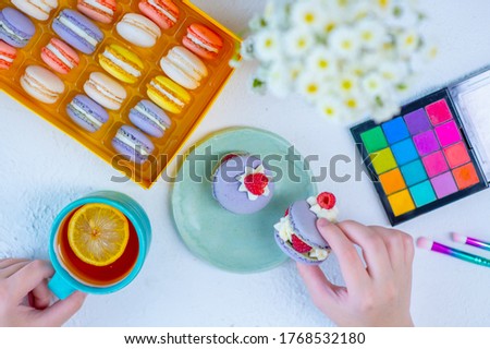 The girl’s hands are holding a cup of tea with lemon and pasta cake, on the table is a full box of cakes and a palette of shadows with tassels. Top view breakfast young girl concept
