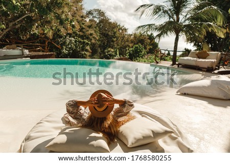 Back view woman wearing straw hat lie on sunbed  and enjoy sun tan near swimming pool with view on ocean and palm trees.  Relaxing summer day, Luxurious tropical vacation concept Royalty-Free Stock Photo #1768580255