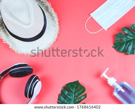 COVID-19 prevention while traveling and new normal lifestyle concept. Top view of surgical  face masks, alcohol  sanitizer gel hat and headphones on  red  background with monstera leaves.