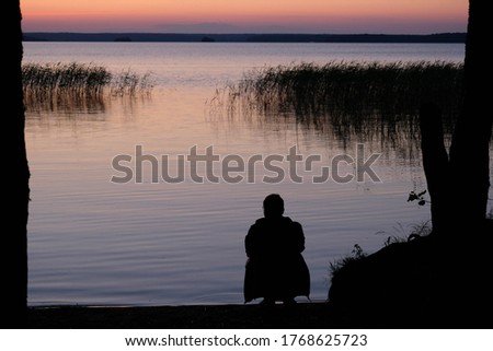 Sunset lake silhouette of a young man looking at the lake, beautiful seascape with sunset. Outdoor summer lake view with dark silhouette on the front. Man looks at the sunset lake. Scenic landscape.