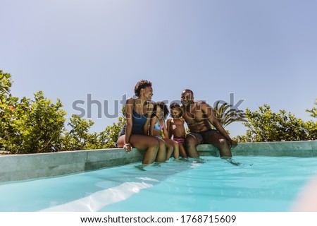 Beautiful young family sitting on the edge of swimming pool with their feet in water. Young couple with their kids enjoying a hot summer day at the poolside.
