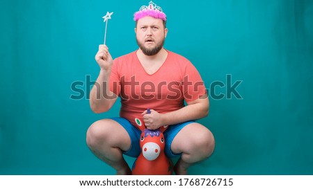 handsome bearded freaky man in a pink T-shirt with a deadema on his head is saddened riding a unicorn with a magic wand in his hand. A funny wizard joke to make and fulfill a wish
