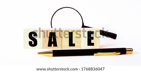 On a light background on wooden blocks between the pen and a magnifying glass the word SALE is written. Financial concept