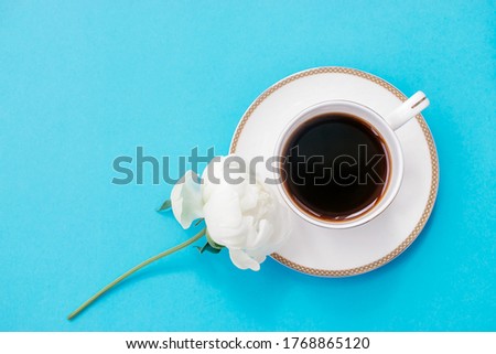 White cup of coffee and white peony flower on a blue background.Summer romantic breakfast.Good morning concept.Selective focus, copy space, top view