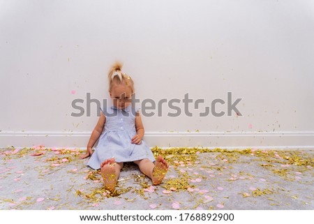 Cute caucasuian blond baby girl sits on a rug with a lot of gold tinsels after celebration holiday, birthday or christmas, feels tired, bare feet, white background, copy space.
