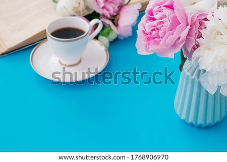 Summer romantic breakfast. Blue vase with peonies, white cup of coffee, open book on a blue background.Good morning concept.Copy space, selective focus with shallow depth of field
