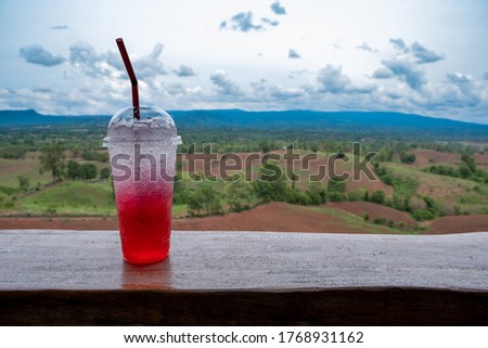 The red drink in a plastic glass is placed on a wooden table, with a mountainous background.