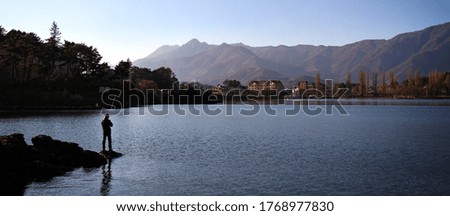 Silhouette of a man standing on rock for fishing in the lake with panorama scenery. Beautiful lake and mountain with blue sky.