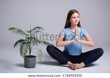 Portrait of yoga trainer girl sitting in lotus position on a white background next to a green flower.