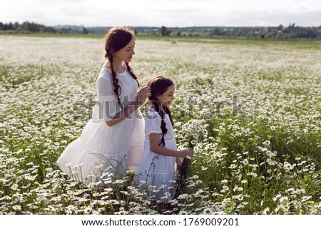 mother with daughter in a white dress and hat stand in a daisy field in summer