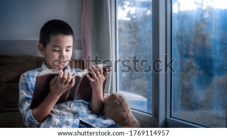 Boy holding and reading Bible on sofa in morning at home. Children's beliefs of christian concept.