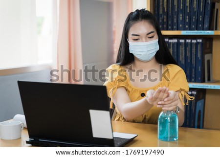 A teenager student wears surgical 
 mask and studies online via laptop during COVID-19 pandemic. University student girl watches online classes and writing a syllabus in notebook. Stock photo.