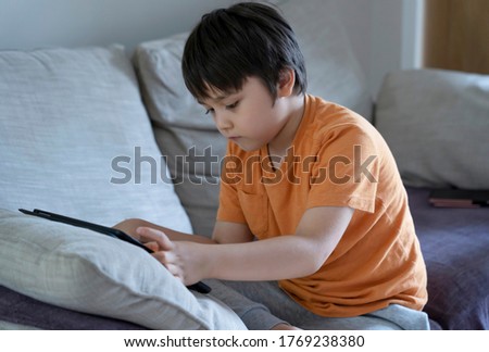 Cropped shot kid sitting on sofa watching cartoons on tablet,6-7 year old boy playing game on touch pad, Cute Kid having fun and relaxing on his own in living room, New normal lifestyle 