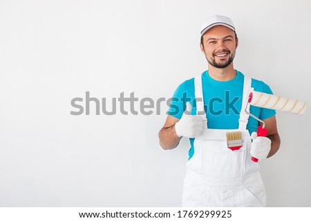 Cheerful man in uniform and with paint roller smiling for camera and gesturing thumb up while recommending renovation works against white background