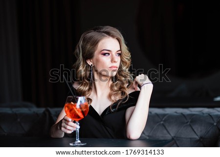 Beautiful blonde, young girl holding an aperol spritz. Aperol cocktail syringe in a glass. Portrait of a young girl in a restaurant. Young girl in a restaurant with a glass on a black background.
