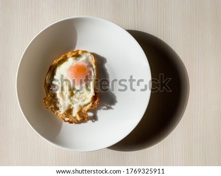 An image of a fried egg placed on a white tile plate and set on the background of a wooden material, which was illuminated in the morning, resulting in light and shadow.