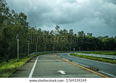 Arrow sign on curved road with green trees in countryside during cloudy day in rain season