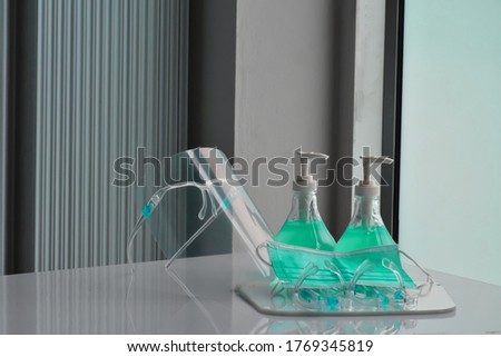 Transparent plastic faceshields, green sanitizers and green medical mask on white table in front of the glass door for wearing on face and rubbing hands, concept for anticoronavirus or covid-19.