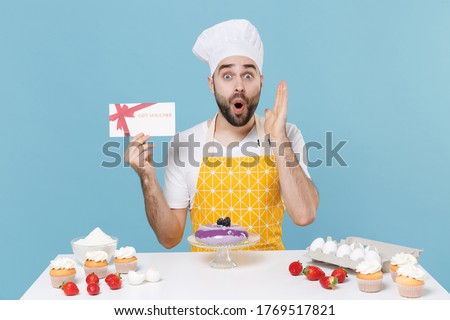 Shocked young bearded male chef or cook baker man in apron white t-shirt toque chefs hat cooking at table isolated on blue background. Cooking food concept. Hold gift certificate, spreading hands