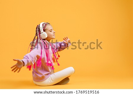 Back view of Adorable child in rounded glasses with pink dreadlocks sitting on floor and listen to music playing in earphones on yellow background. Cute kid listening to music in headset, copy space.