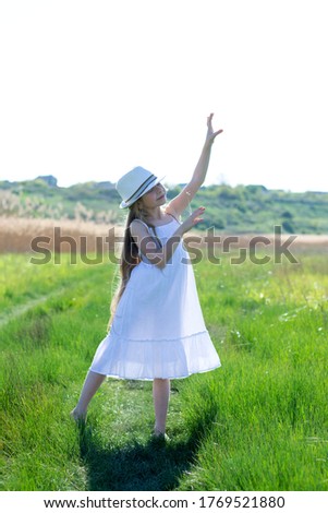 8-year-old girl with long blonde hair in a white sundress and white hat dances barefoot among the lush spring grass