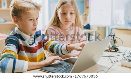 Brilliant Schoolgirl and Schoolboy Talk and Use Laptop to Program Software for Robotics Engineering Class. Elementary School Science Classroom with Children Working on Technology. STEM Education Royalty-Free Stock Photo #1769673836