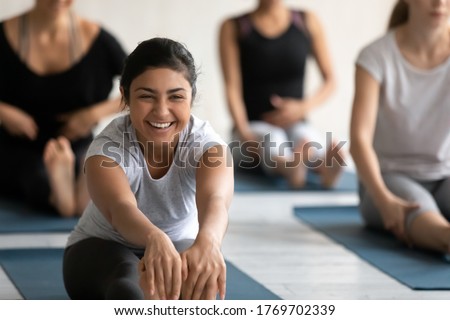 Happy young indian woman excited by flexibility progress in seated forward bend position, enjoying group yoga class indoors. Smiling beautiful fit biracial lady practicing Paschimottanasana in studio. Royalty-Free Stock Photo #1769702339