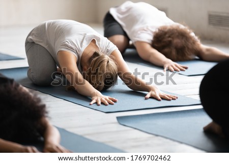 Focus on young female beginner lying on floor mat in balasana pose, relaxing muscles after intensive fitness pilates or stretching body after workout at group yoga class in modern studio interior. Royalty-Free Stock Photo #1769702462